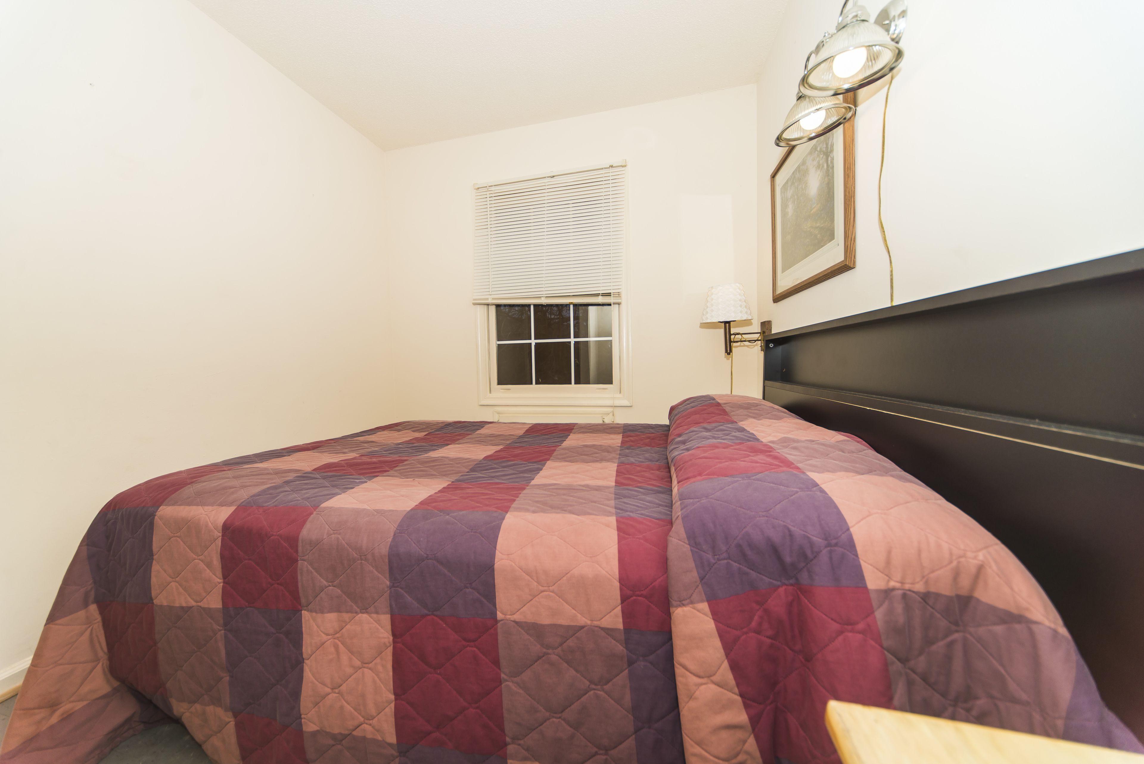 A bedroom with a bed covered in a quilted blanket, a window with blinds, and a wall-mounted lamp.