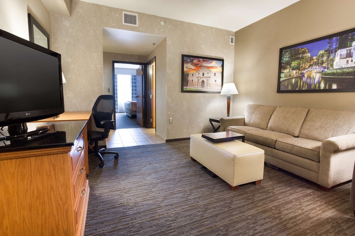 Une chambre d’hôtel équipée d’un téléviseur à écran plat, d’un canapé beige, d’un pouf blanc et d’un bureau avec une lampe.