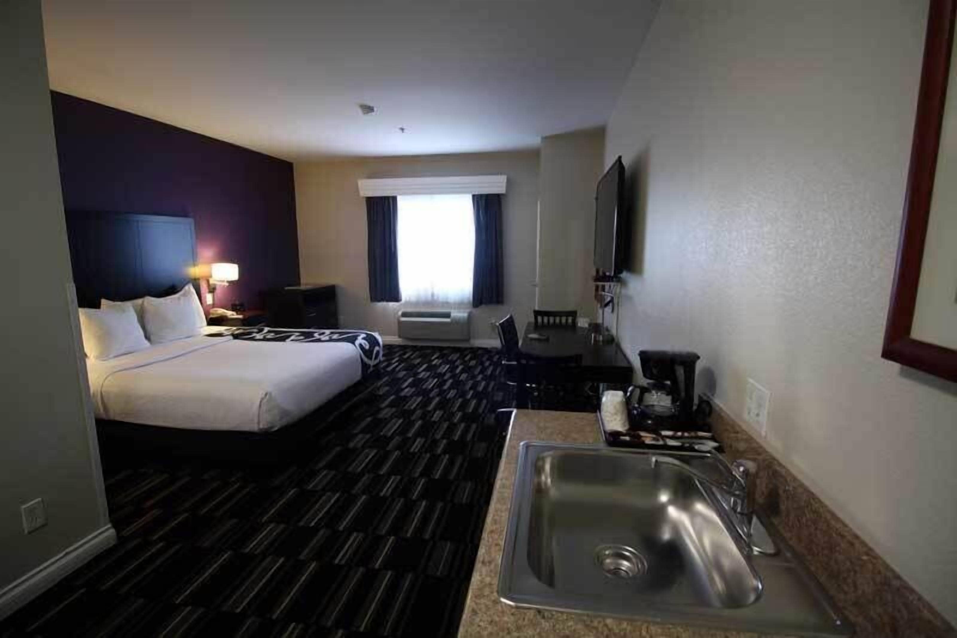 A hotel room with a bed, a desk, a chair, a TV, a window with curtains, a sink, and a mirror.