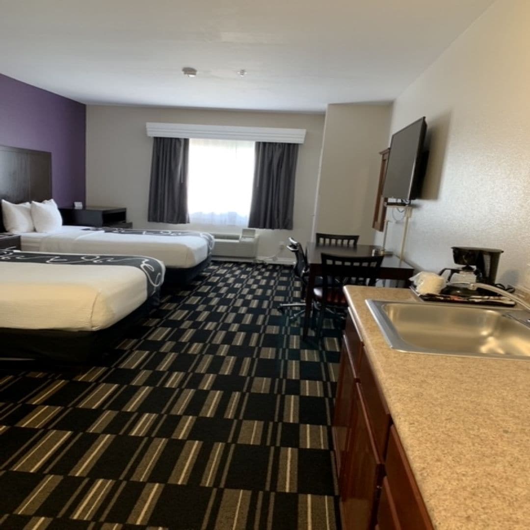 Habitación de hotel con dos camas, kitchenette, zona de comedor y un ventanal con cortinas.