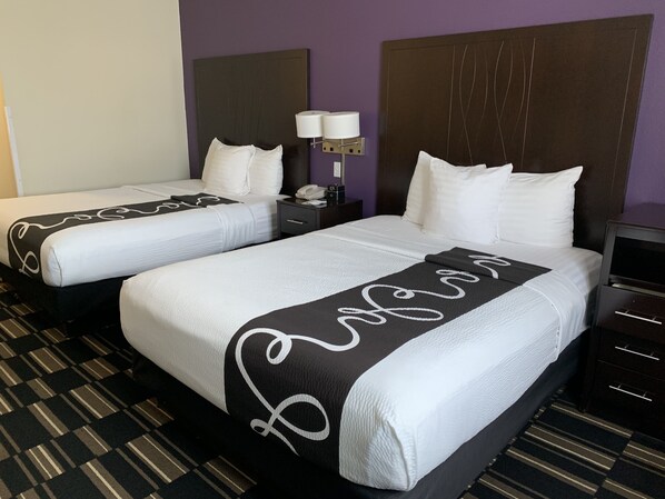 A hotel room with two beds, each with white linens and a black and white patterned throw. There is a nightstand with a lamp and a phone between the beds. The room has a purple wall and a checkered floor.