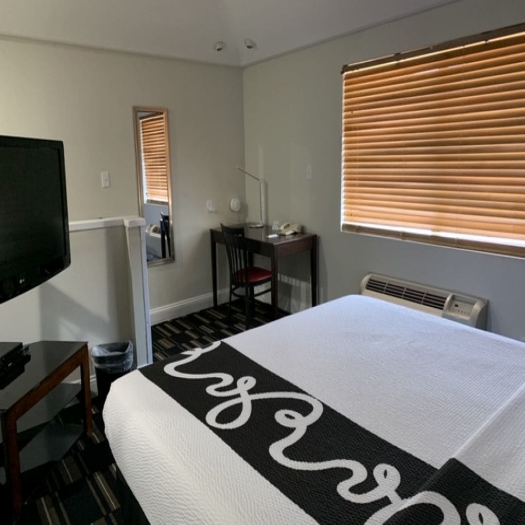 Habitación de hotel con una cama grande, un escritorio, una silla, un televisor y una ventana con persianas.