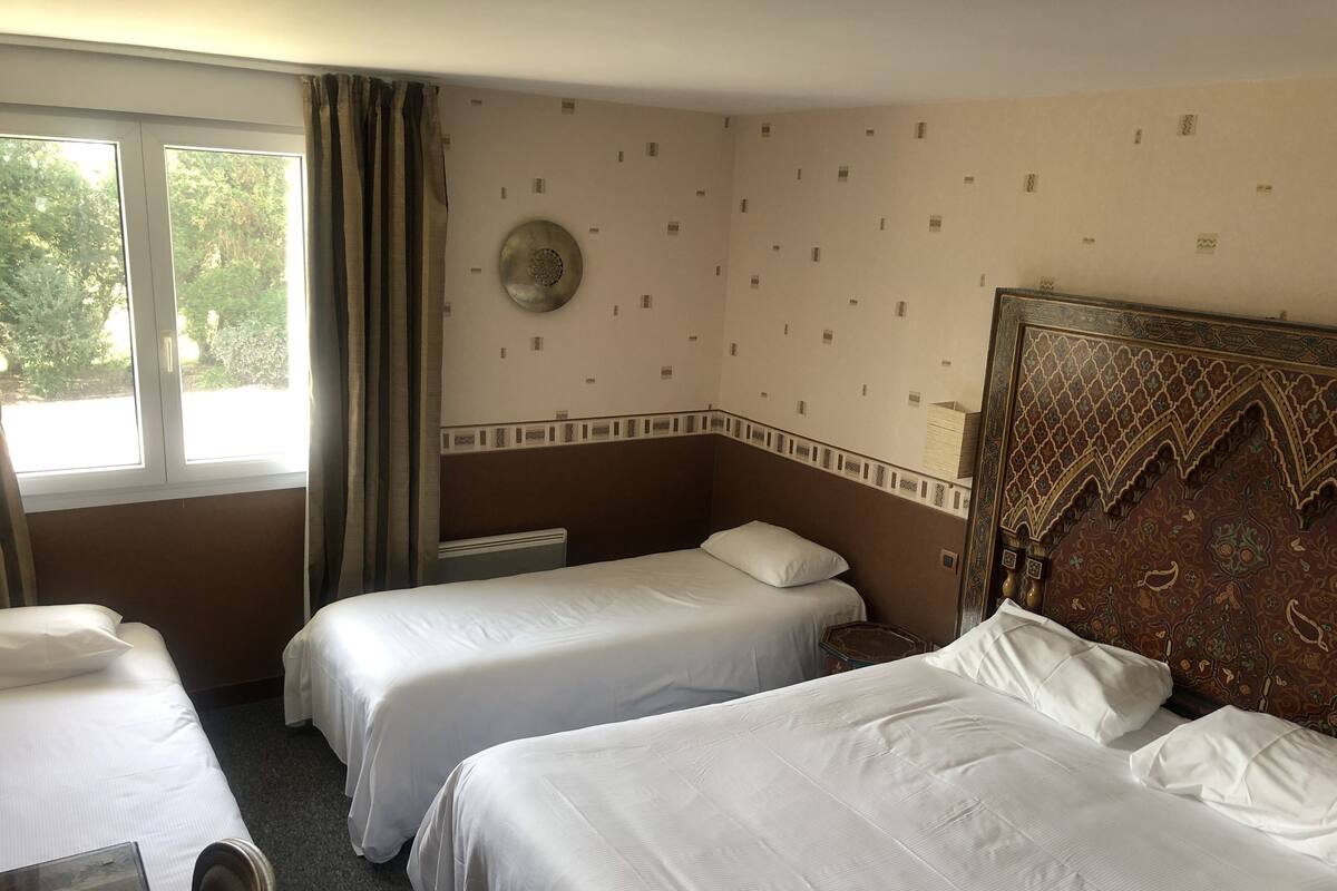 Une chambre d’hôtel avec deux lits simples, une grande fenêtre avec des rideaux, une horloge murale et une tête de lit décorative.