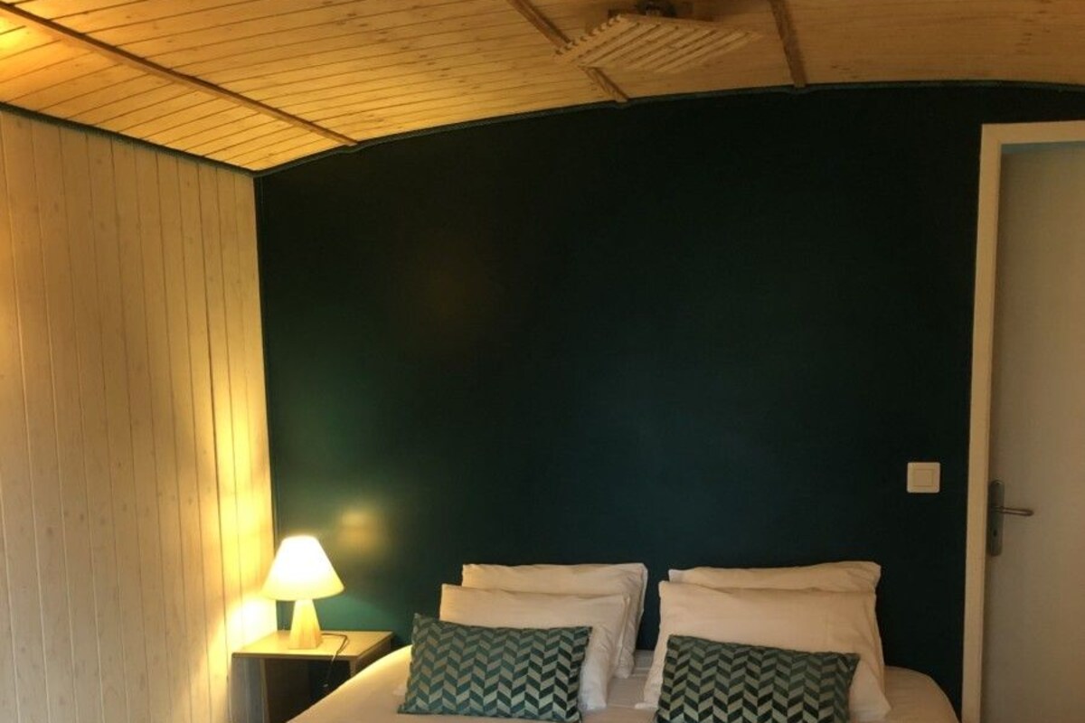 A bedroom with a wooden ceiling, a bed with white bedding, two pillows with a green pattern, and a bedside lamp.
