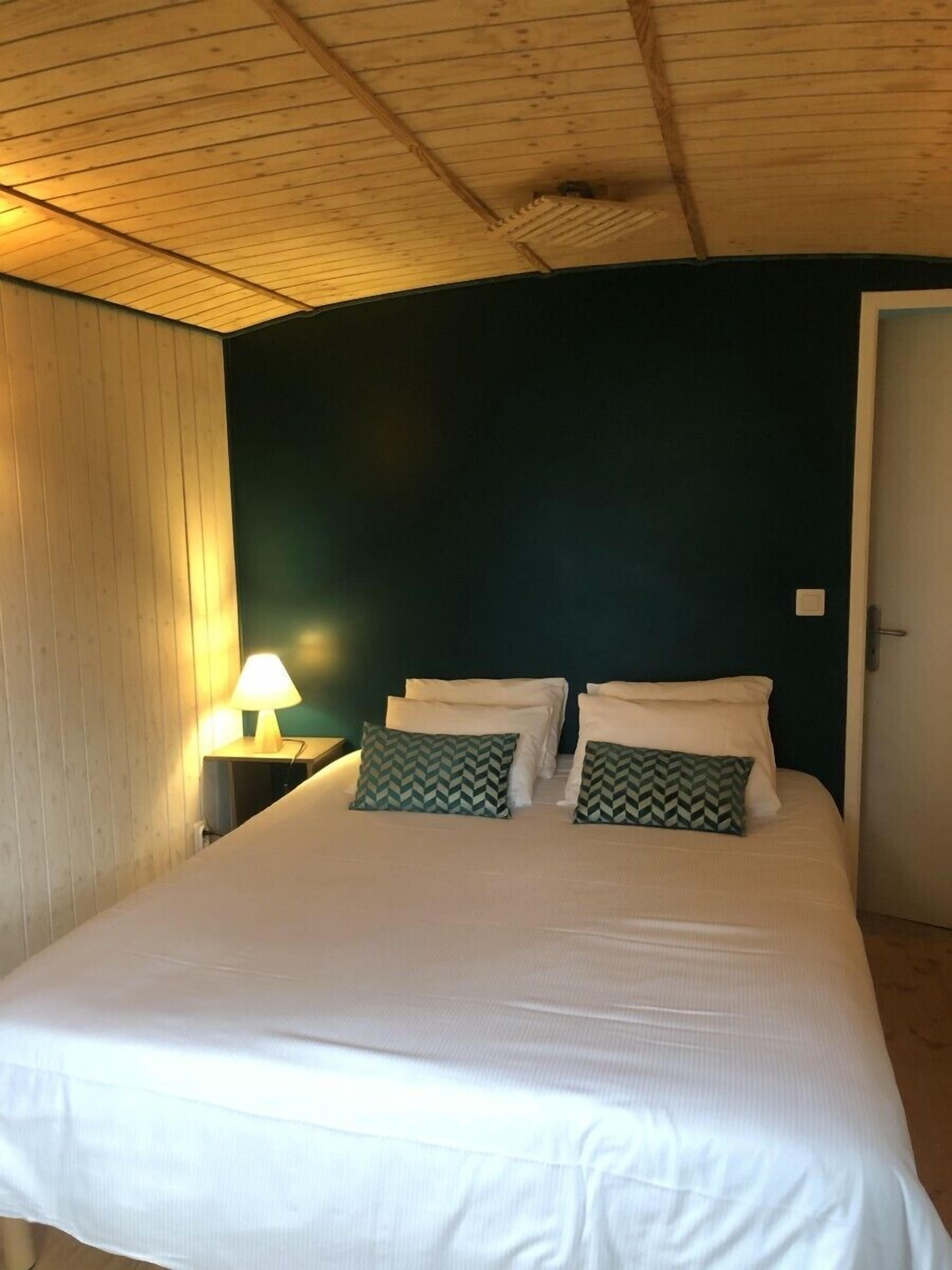 Une chambre avec un plafond en bois, un lit avec une literie blanche, deux oreillers à motifs verts et une lampe de chevet.