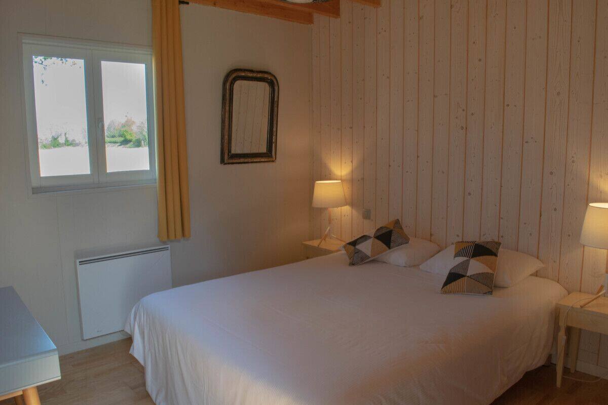 A bedroom with a bed, two bedside lamps, a window with curtains, a radiator, and a wooden wall paneling.