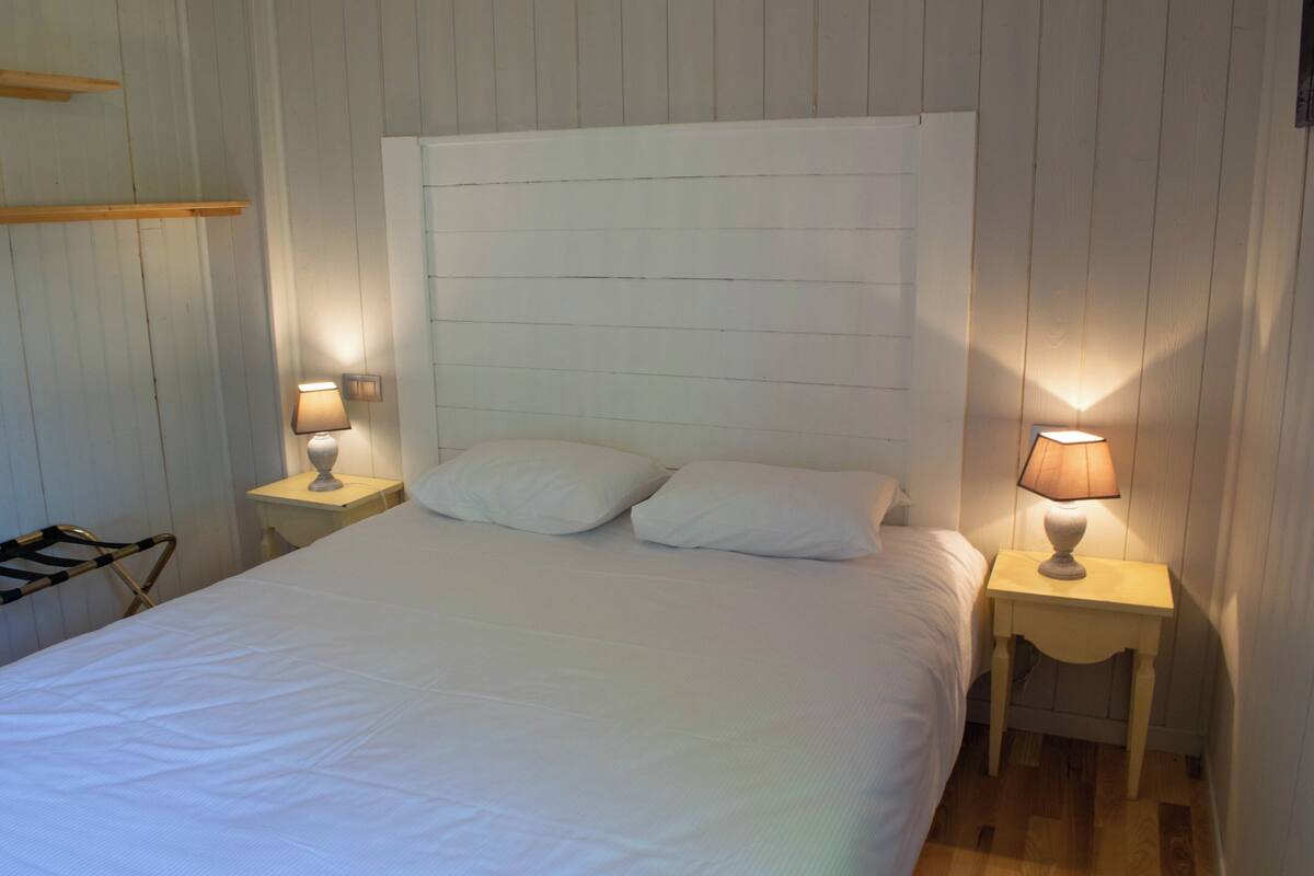 Un lit bien fait, avec du linge de lit blanc, dans une pièce aux murs recouverts de lambris en bois. On y trouve deux tables de chevet avec des lampes.