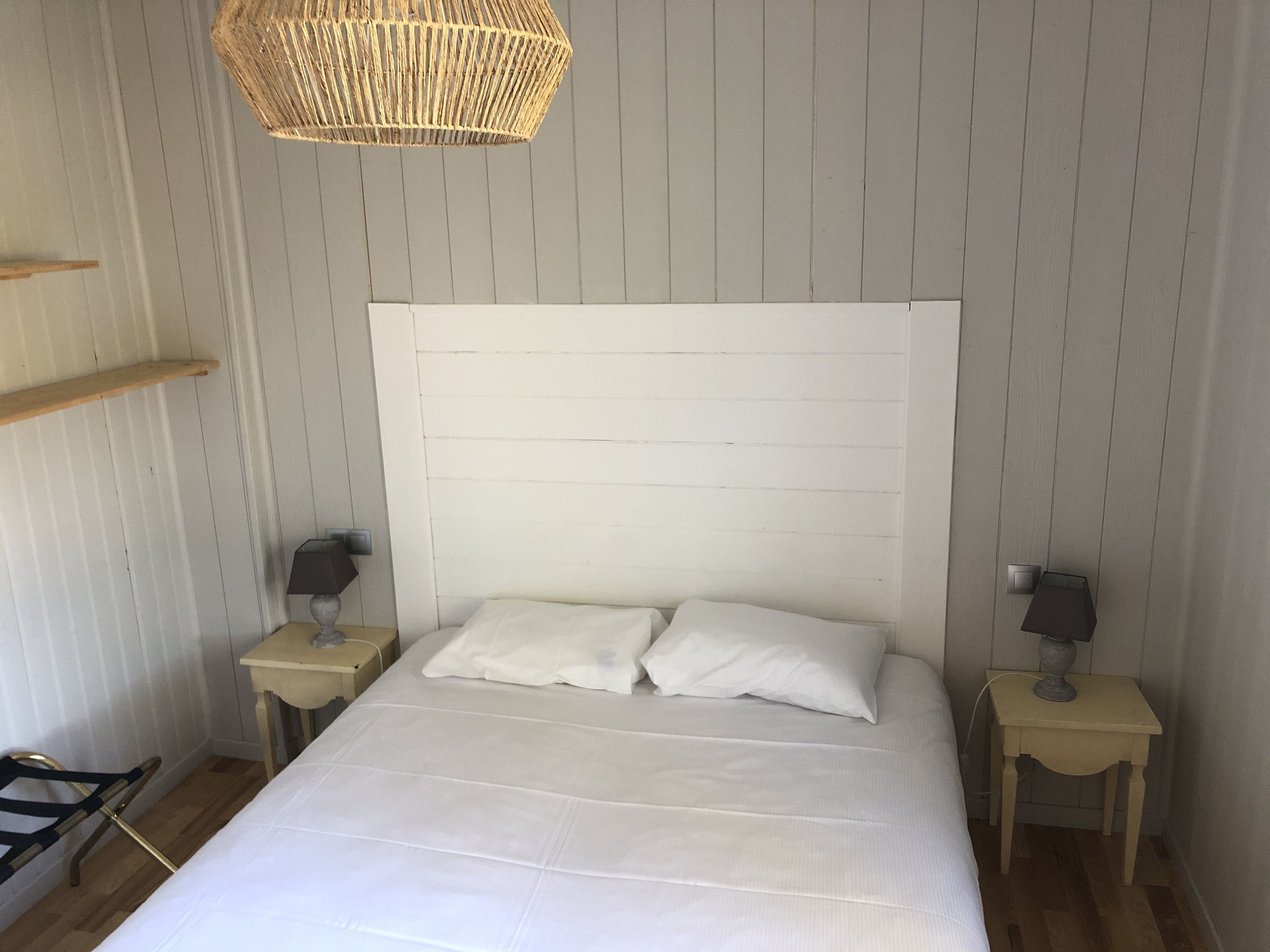 A bedroom with a large bed, two bedside tables, a hanging light fixture, and a wooden shelf.