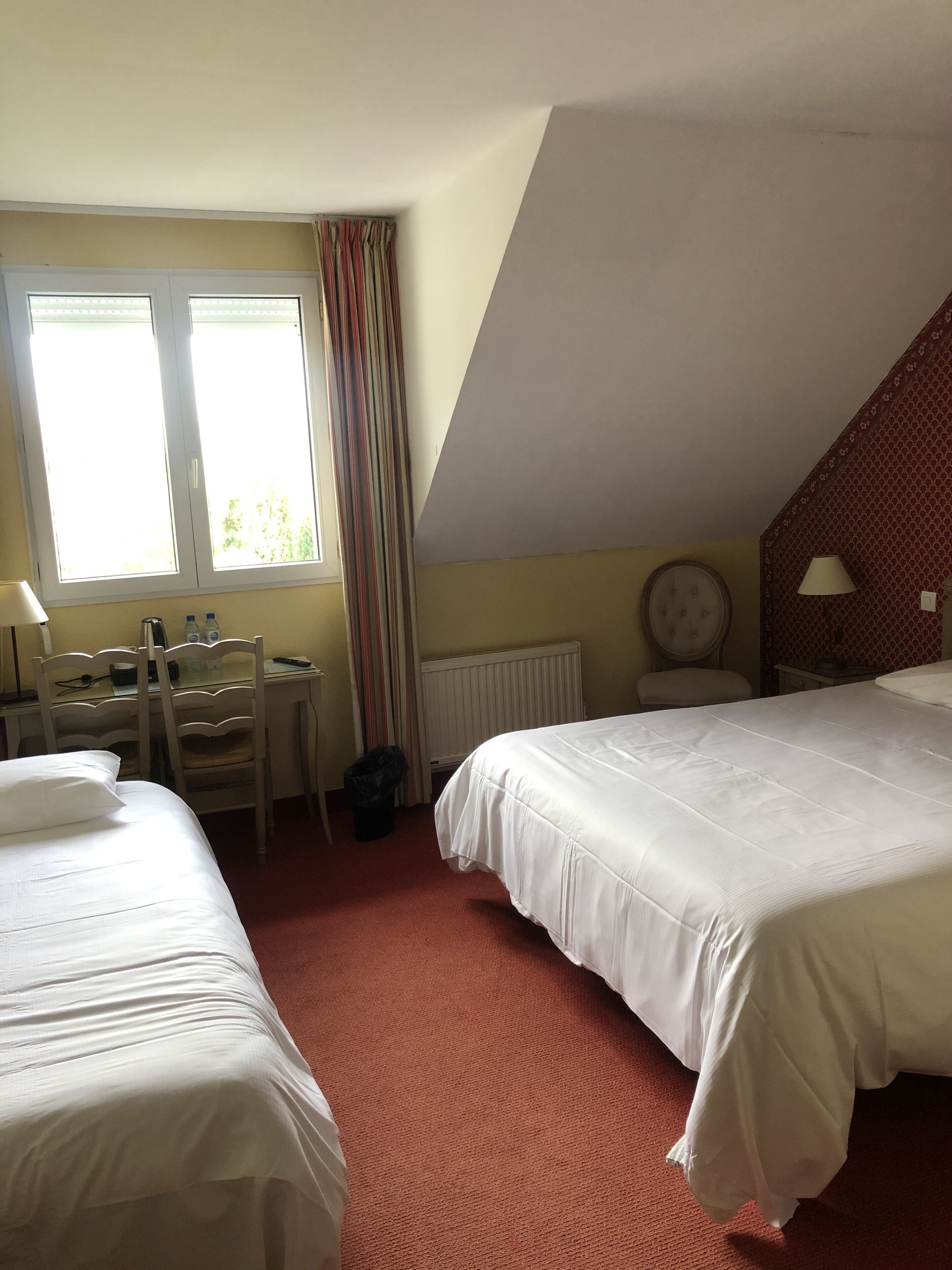A hotel room with two beds, a desk with a chair, a radiator, and a window with curtains.