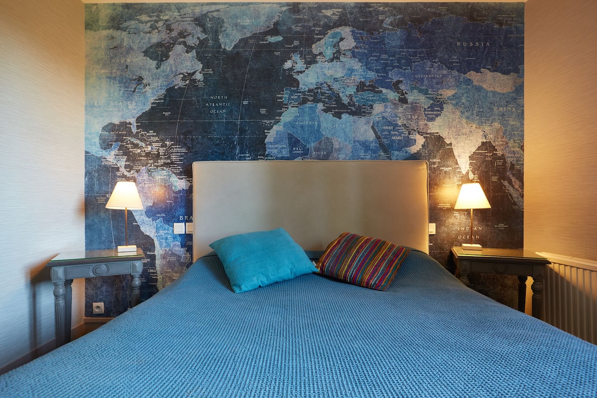 Un lit avec une couvre-lit bleue et deux oreillers, une carte du monde accrochée au mur et deux lampes de chevet.