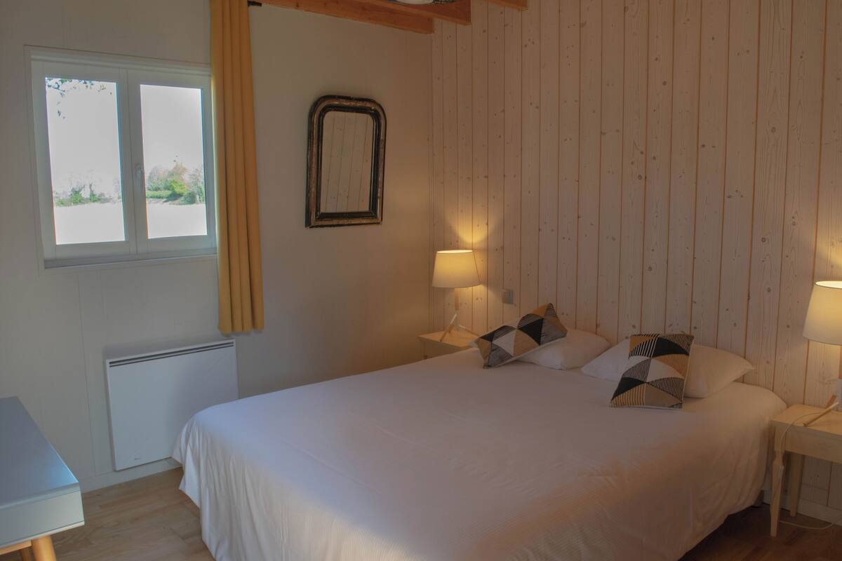 Une chambre à coucher avec un lit, deux lampes de chevet, une fenêtre avec des rideaux, un radiateur et un panneau mural en bois.