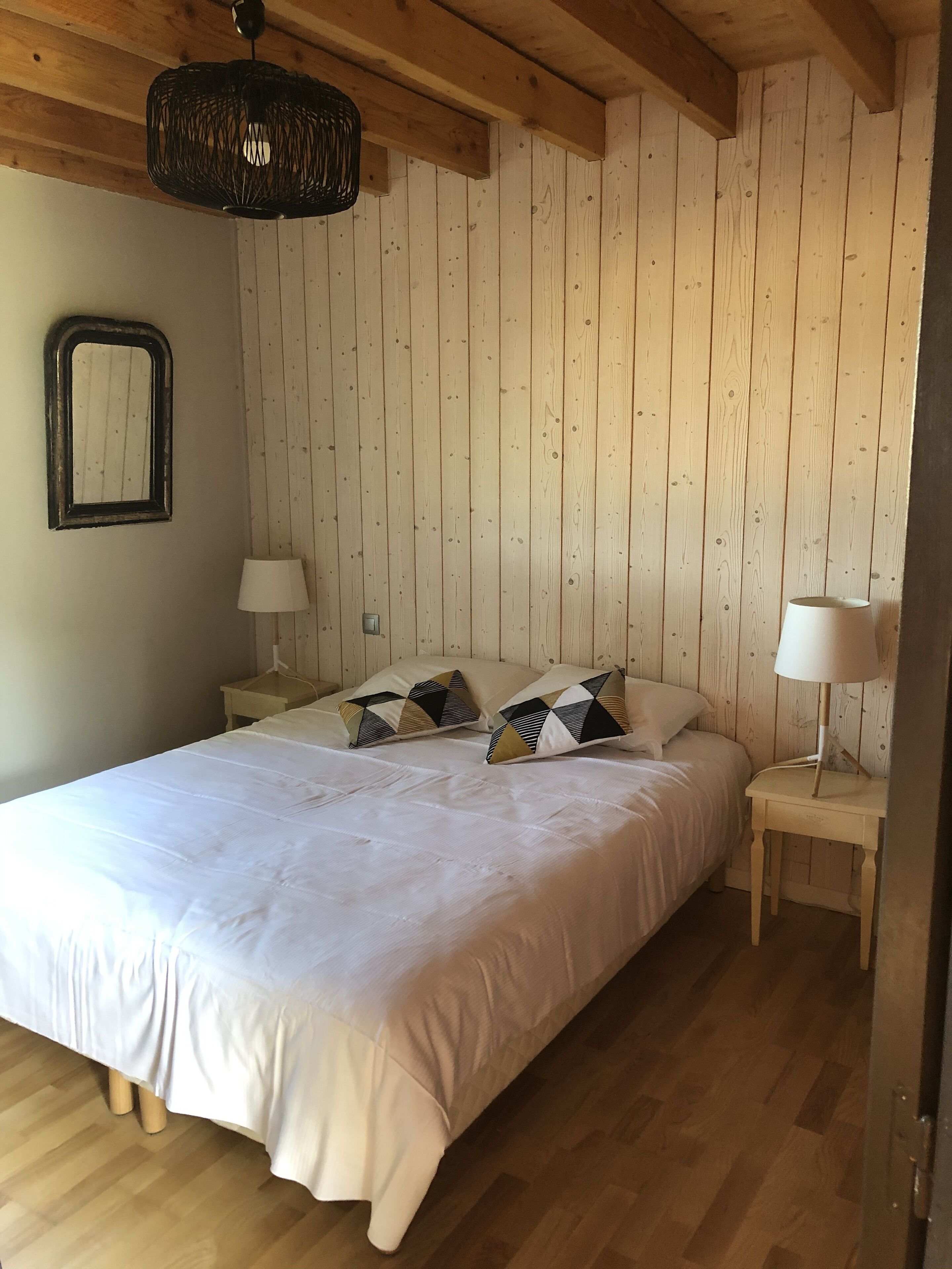 Un dormitorio con piso de madera, una cama con ropa blanca, dos mesitas de noche con lámparas y un candelabro colgante.