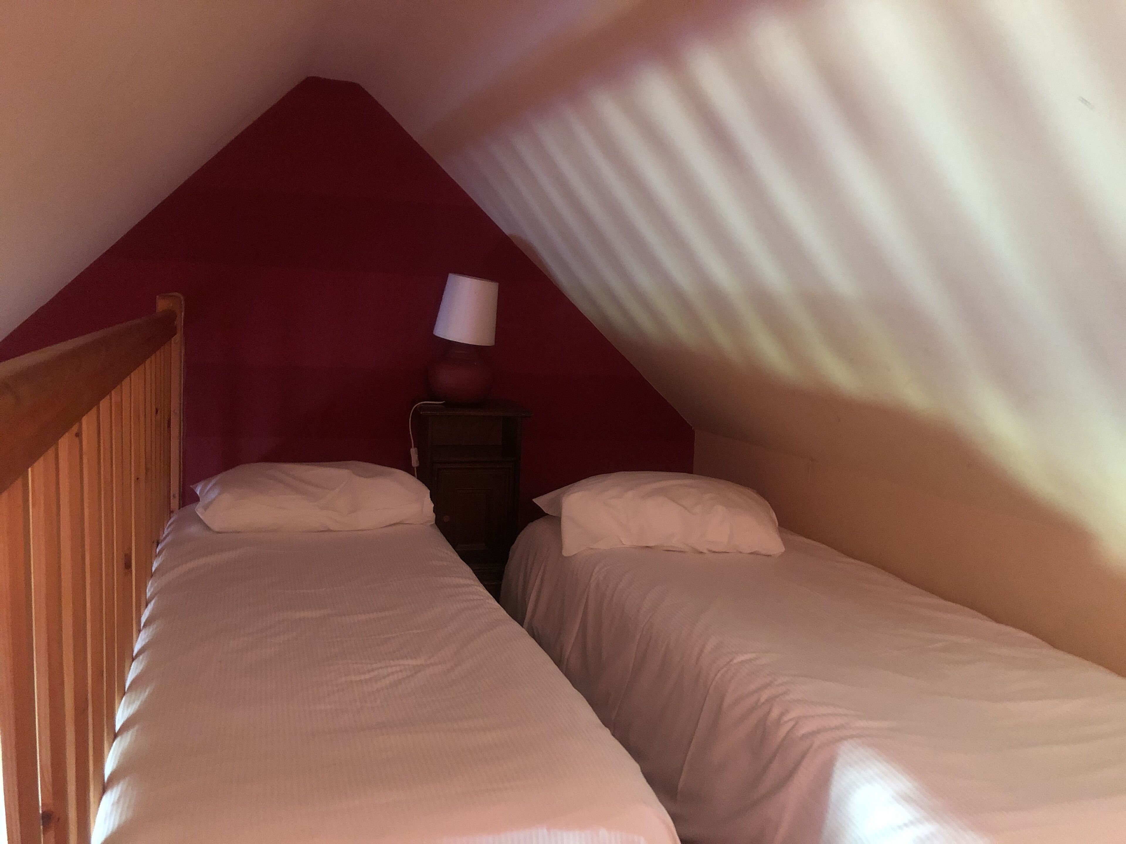 Une petite pièce peu éclairée, avec deux lits simples, une tête de lit en bois, une table de chevet avec une lampe et un plafond en pente.