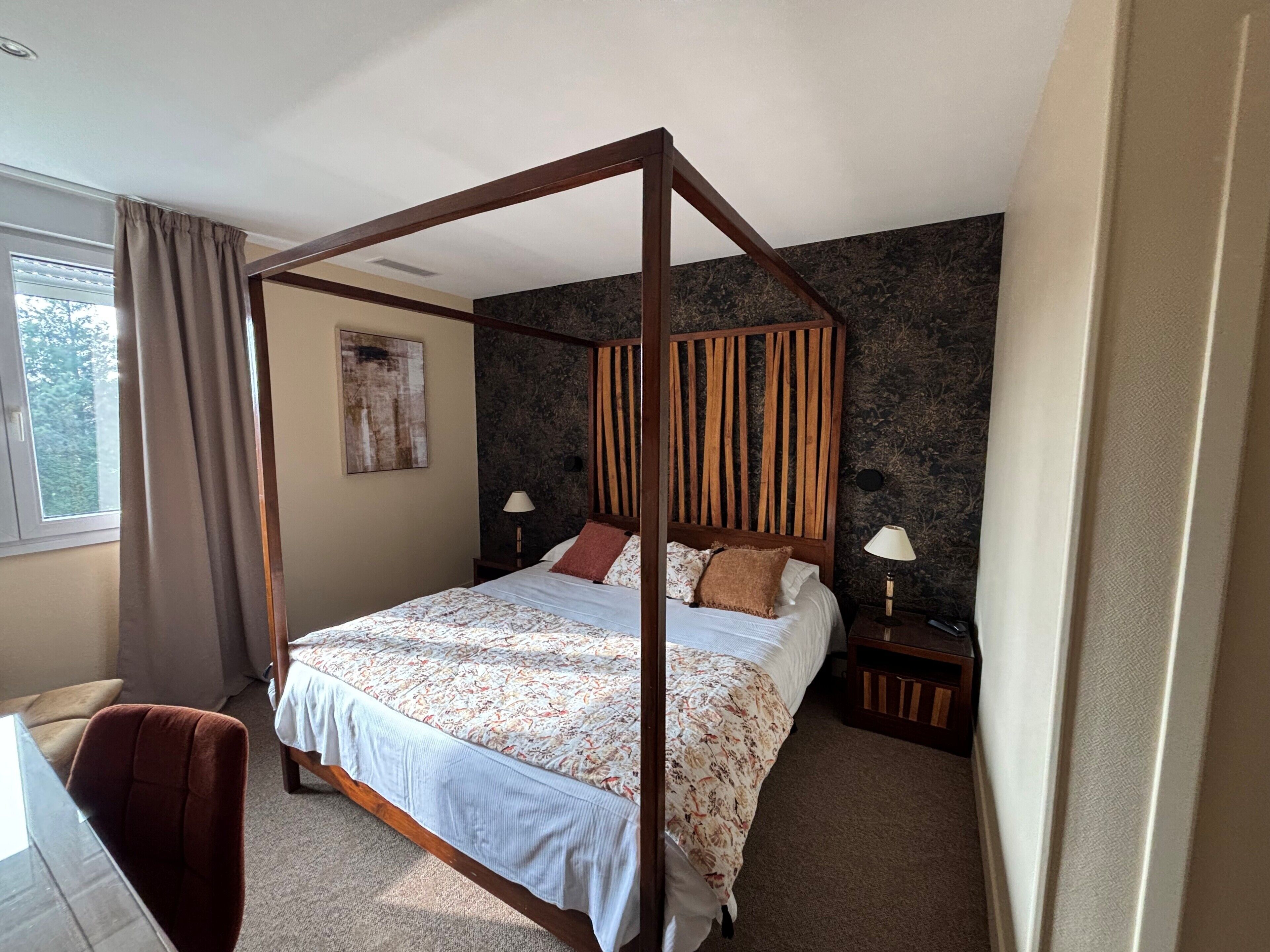Ein Bett mit Baldachin, ein Kopfteil, zwei Nachttische mit Lampen, ein Stuhl, ein Fenster mit Vorhängen und eine Wand mit strukturierter Oberfläche.