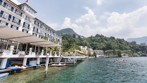 Hotel frente al agua con balcones, un muelle con barcos y montañas al fondo.