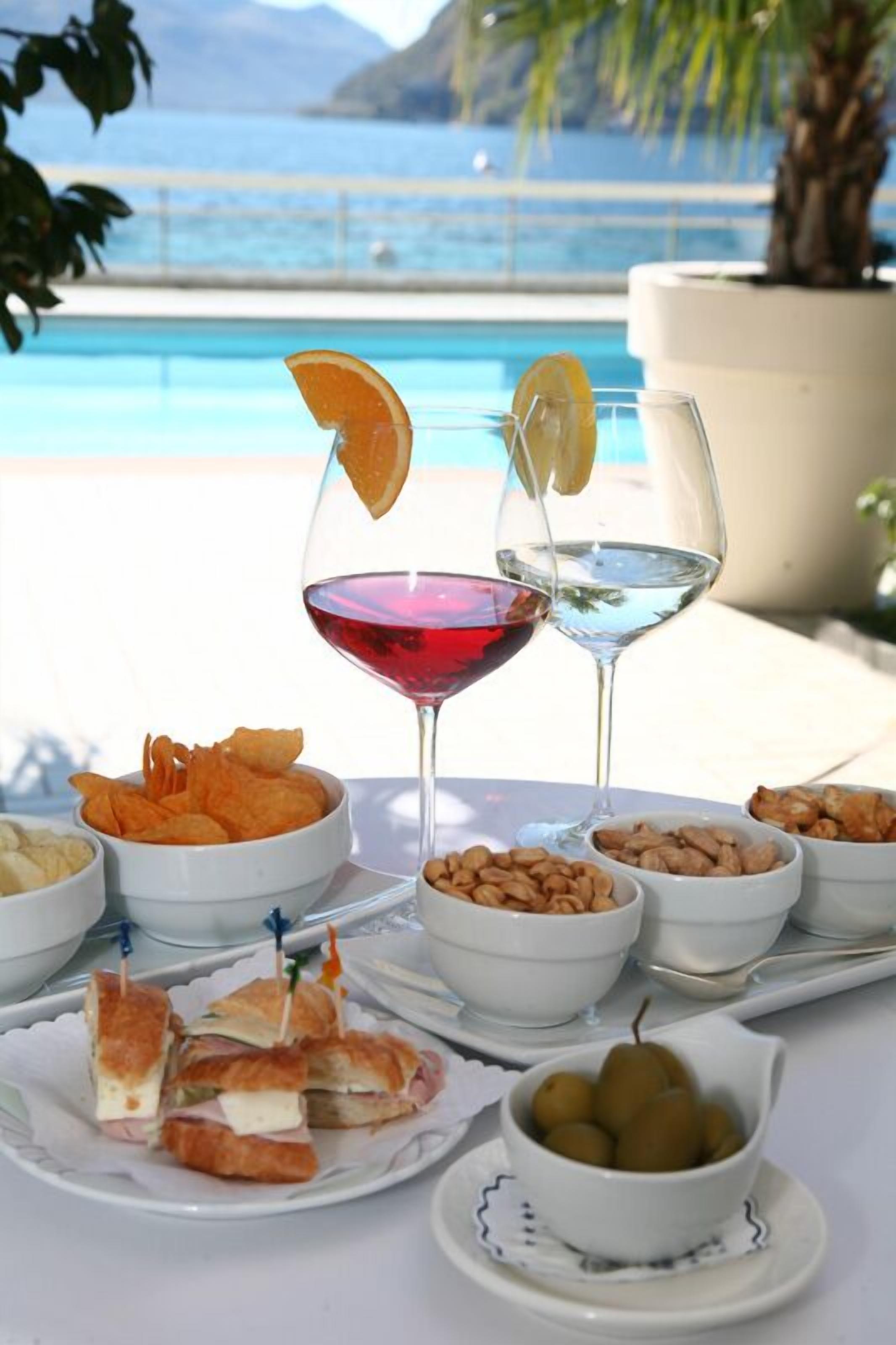 A table with a variety of snacks, including chips, nuts, and fruit, alongside two glasses of red and white wine, set against a backdrop of a pool and mountains.