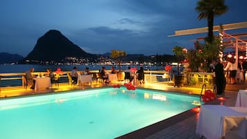 A poolside dining area with tables and chairs, overlooking a body of water and a mountain.