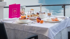 Una mesa dispuesta con alimentos para el desayuno, un menú rosa y vista a un mar tranquilo y una montaña.