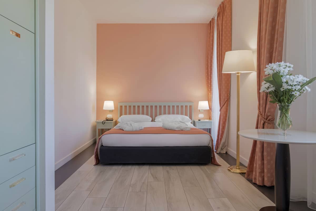 A hotel room with a bed, nightstands, a lamp, a vase with flowers, and a wardrobe.