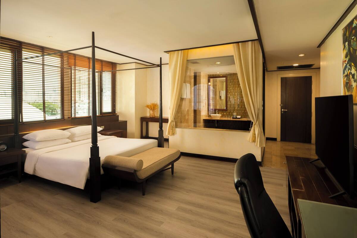 Une chambre d’hôtel moderne avec un lit à baldaquin, un coin salon avec un banc, une télévision et une grande fenêtre équipée de stores.