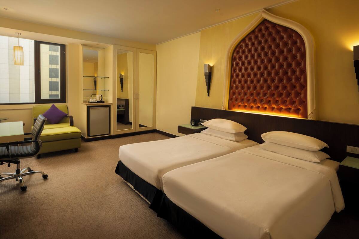 Une chambre d’hôtel avec deux lits, un bureau avec une chaise, une petite kitchenette et une grande fenêtre en arc de cercle.
