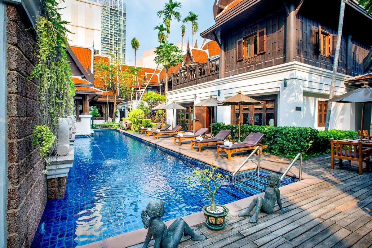 Une piscine entourée d’une terrasse en bois et de verdure, avec des chaises longues et une statue.