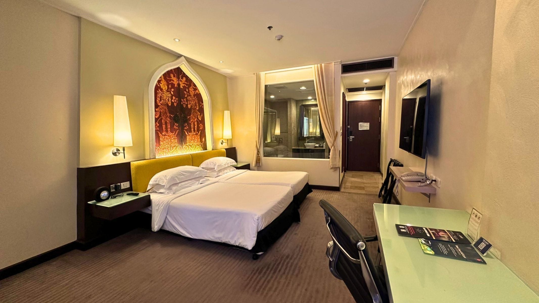 Habitación de hotel con cama, lámparas de noche, escritorio con silla, televisor y baño visible a través de una puerta de cristal.
