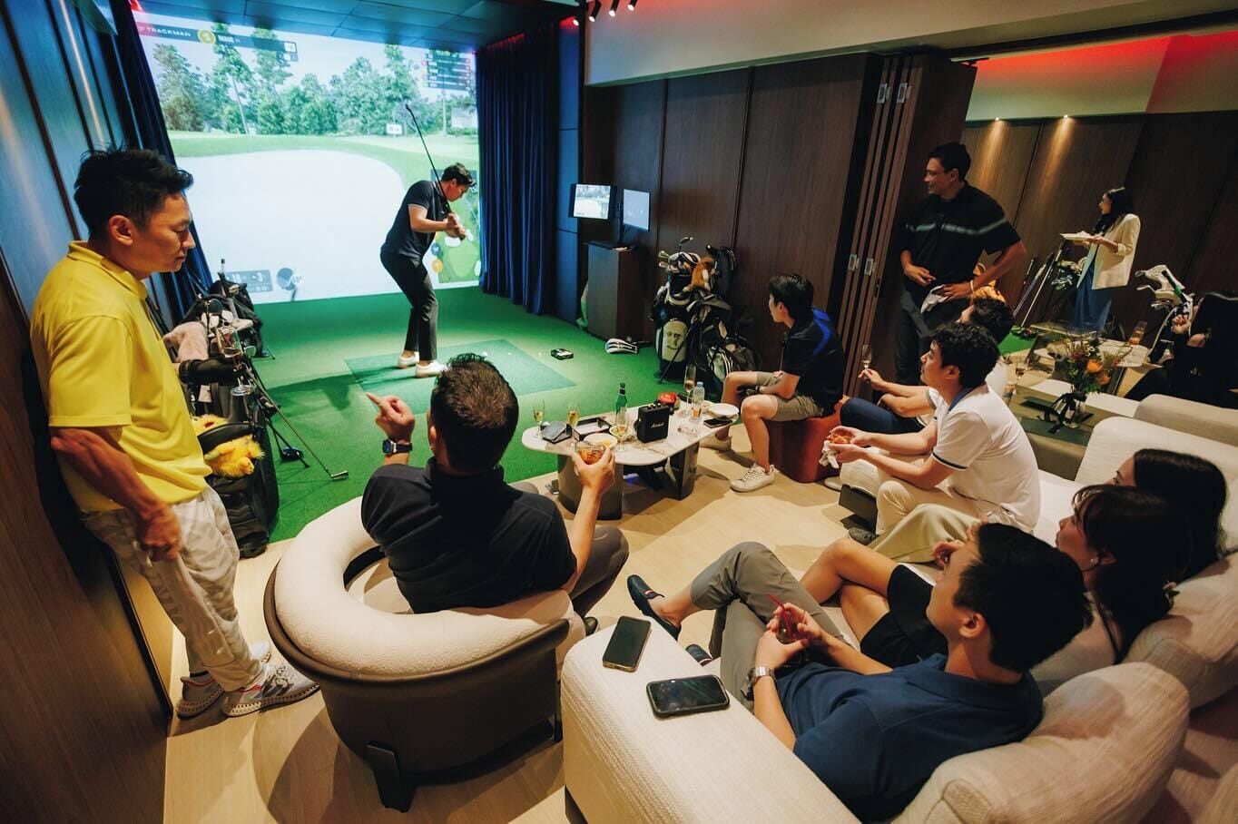 Sala de simulación de golf con pantalla verde y proyección de campo de golf, rodeada de invitados sentados y un golfista practicando.