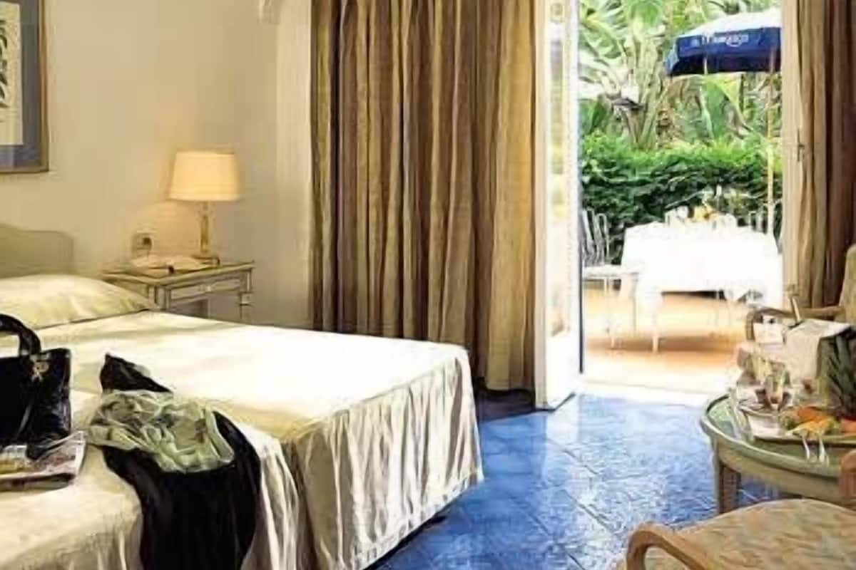 Hotellihuone, jossa on sänky, yöpöytä, lamppu ja näkymä ulkoilmaravintolaan.
