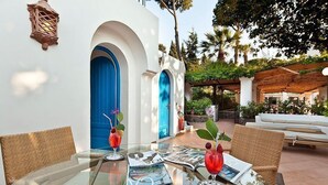 A glass table with magazines, two red vases with flowers, and wicker chairs in a sunny outdoor area with white walls and blue doors. - Grand Hotel Il Moresco & Spa (Ischia)