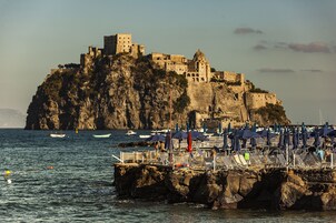 Vista costera con un castillo histórico en un acantilado, un muelle con sombrillas y sillas, y barcos en el agua.