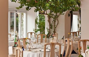 A dining area with round tables, wooden chairs, and a large tree with green leaves.