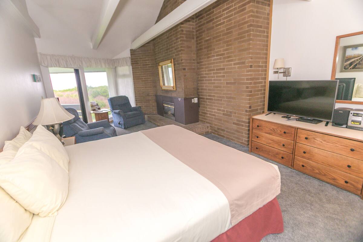 Quarto de hotel com uma cama grande, televisão, lareira e secretária com abajur.