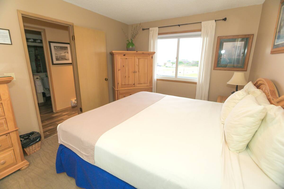Quarto com cama, guarda-roupa de madeira, janela com cortinas e um quadro na parede.
