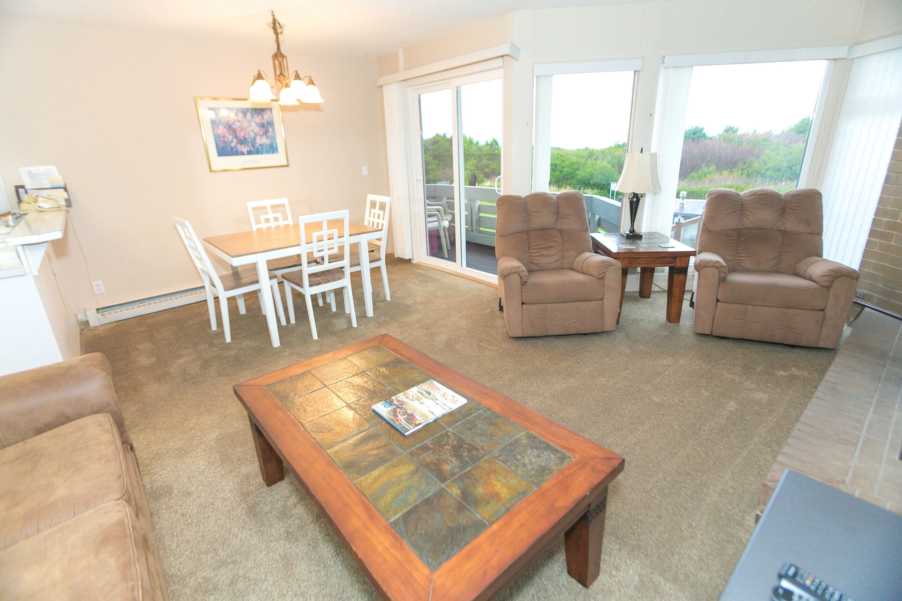 A living room with a coffee table, two armchairs, a dining table with chairs, a TV, and a sliding glass door leading to a balcony.