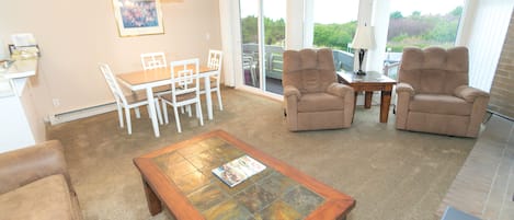 A living room with a coffee table, two armchairs, a dining table with chairs, a TV, and a sliding glass door leading to a balcony.