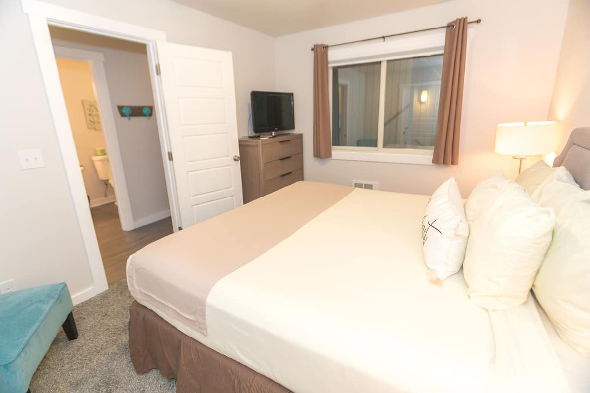 Quarto com cama, televisão sobre cômoda, janela com cortinas e banheiro visível por uma porta aberta.