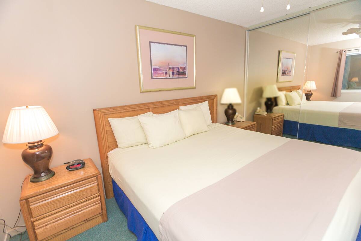 Quarto de hotel com cama, luminárias de cabeceira, mesa de cabeceira, gaveta, quadro pendurado na parede e espelho refletindo o quarto.