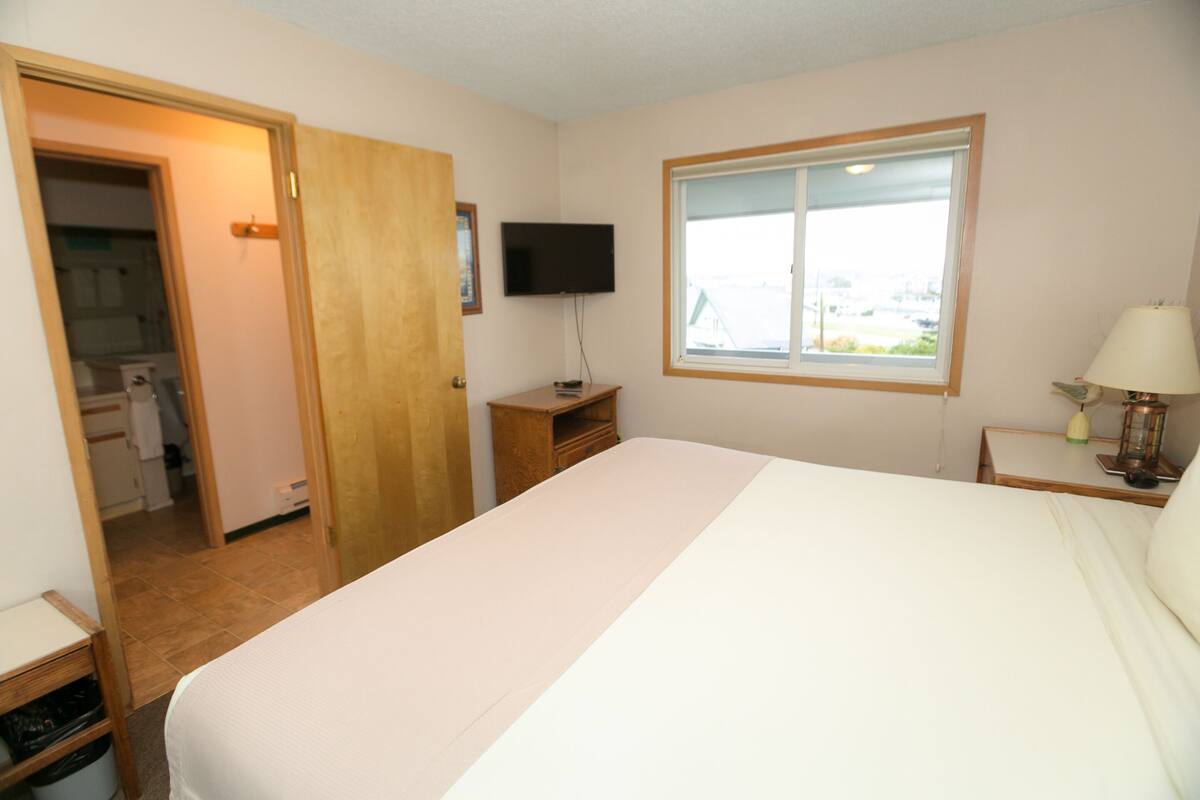 Quarto de hotel com cama, televisão, janela, mesa de cabeceira e porta de madeira que dá para um banheiro.
