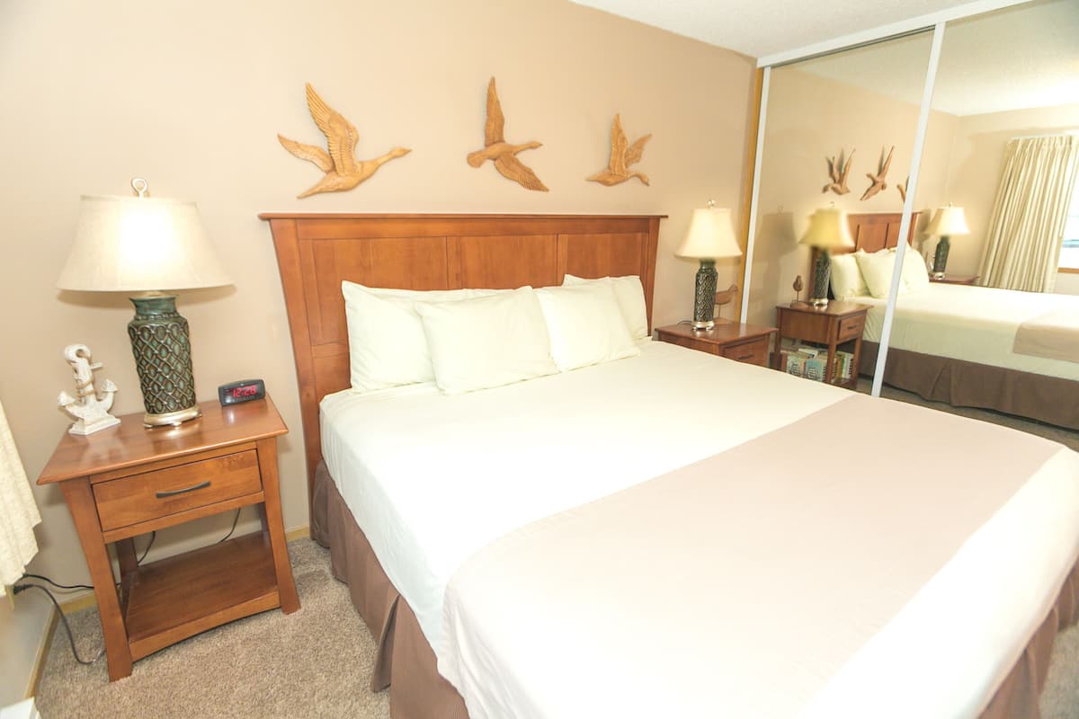 Quarto de hotel com uma cama grande, mesas de cabeceira de madeira com abajures, um espelho e uma pintura de pássaros voando.
