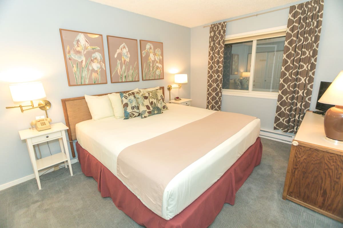 Quarto de hotel com cama grande, duas mesas de cabeceira, abajur e janela com cortinas estampadas.