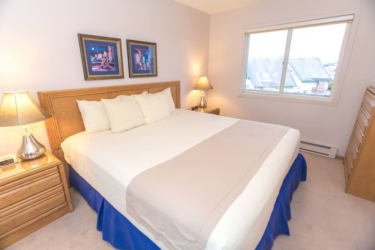 Quarto de hotel com uma cama grande, duas mesas de cabeceira, uma janela e duas imagens emolduradas na parede.