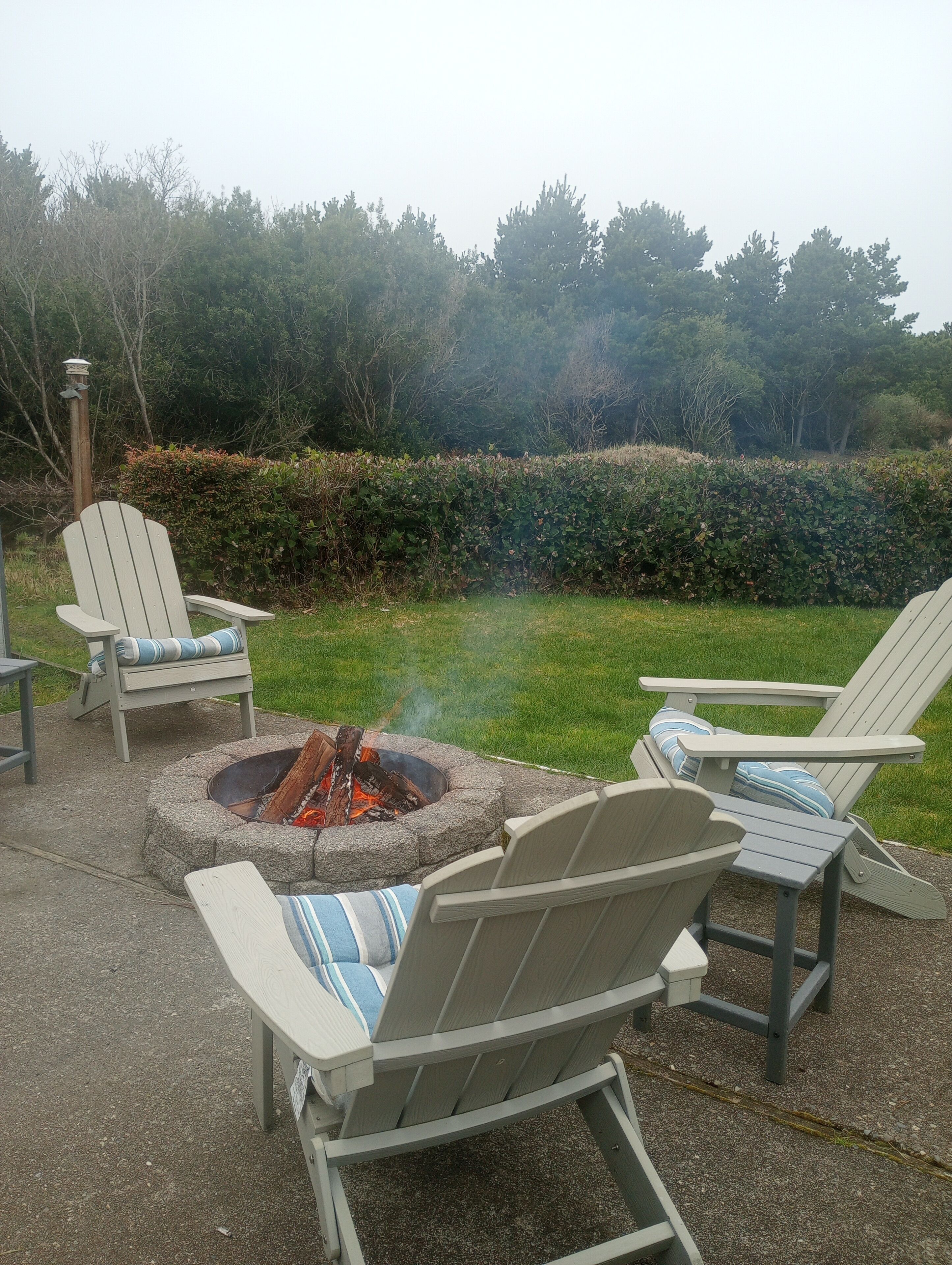 A fire pit with chairs around it, a wooden post, and a hedge in the background.