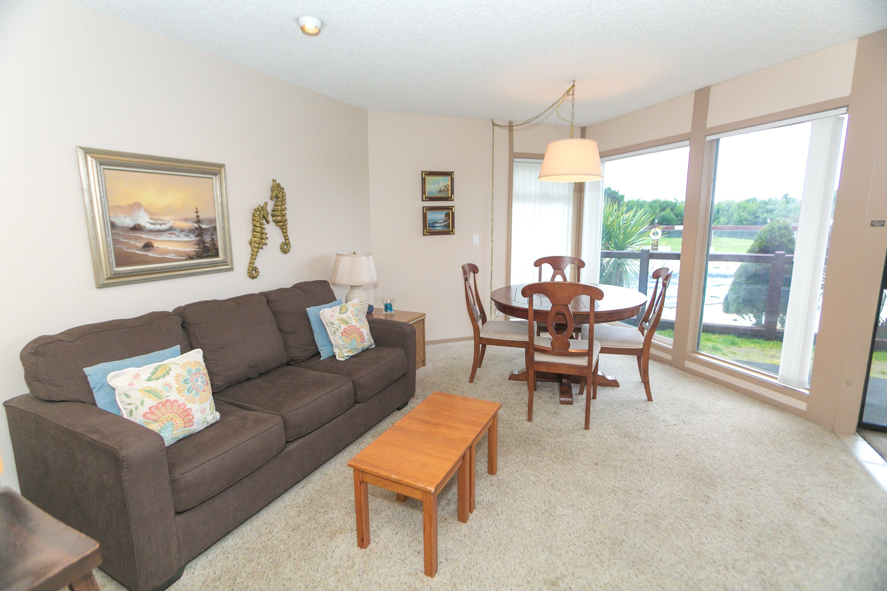 A living room with a brown sofa, a wooden coffee table, a dining table with chairs, and a large window offering a view of the outdoors.