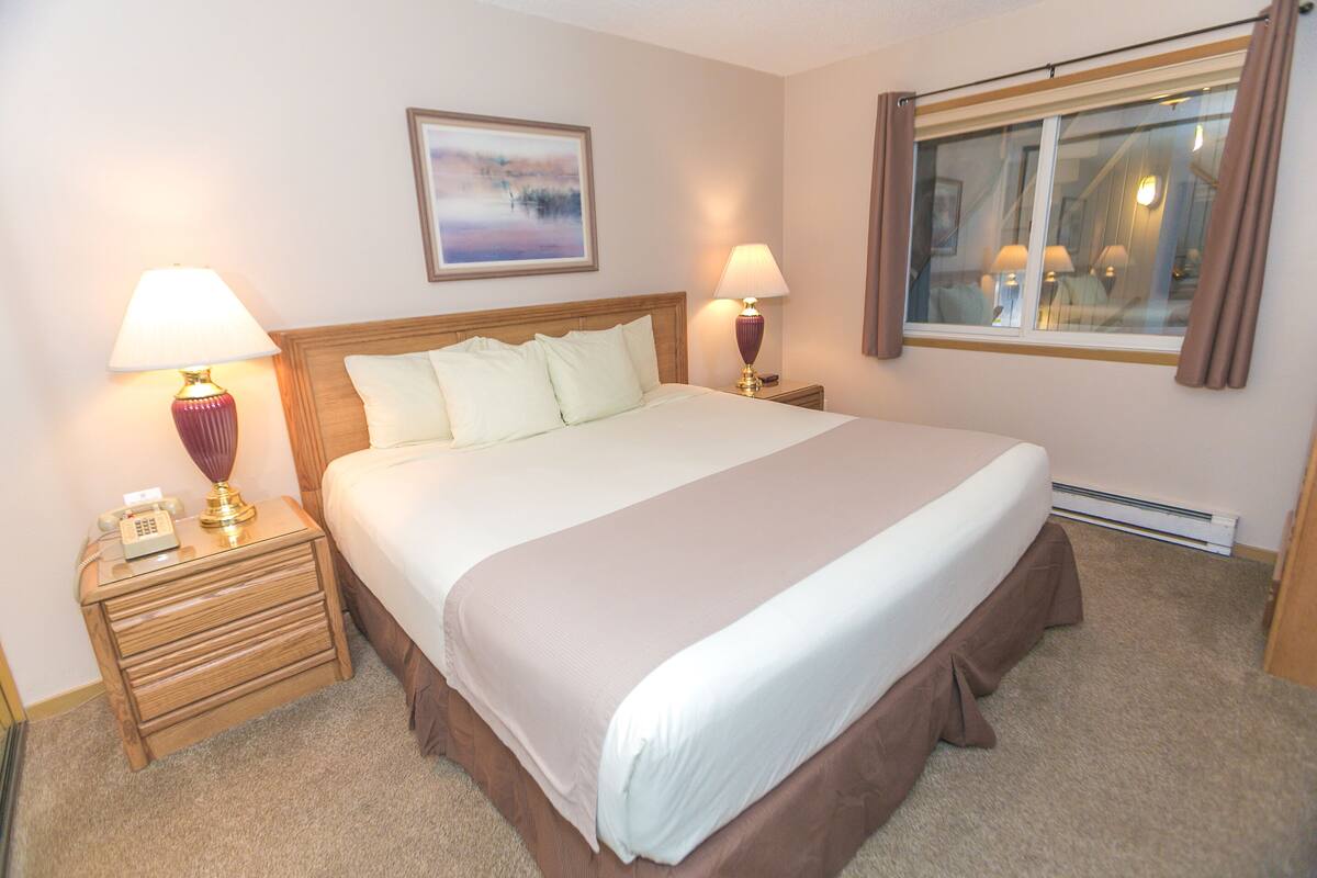 Cama bem arrumada com cabeceira, duas mesas de cabeceira com abajures, um quadro na parede e uma janela com cortinas.