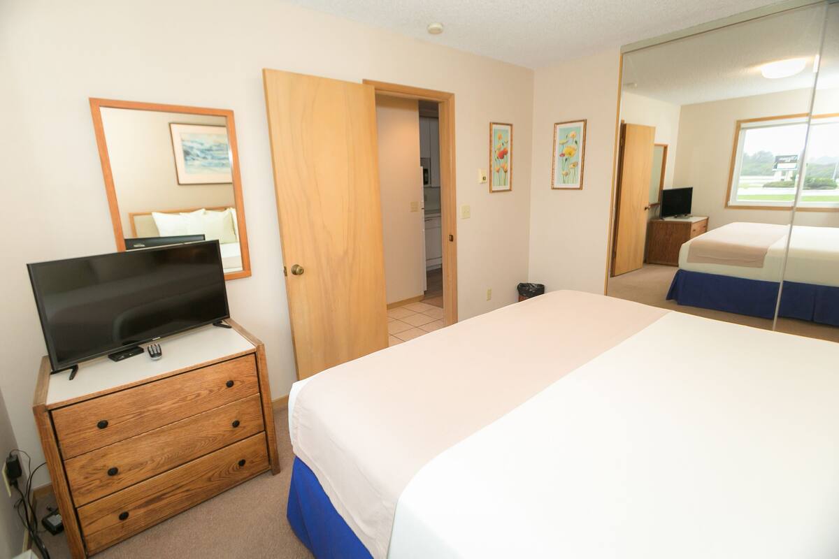 Quarto de hotel com cama, televisão em cômoda de madeira, espelho e porta para o banheiro.
