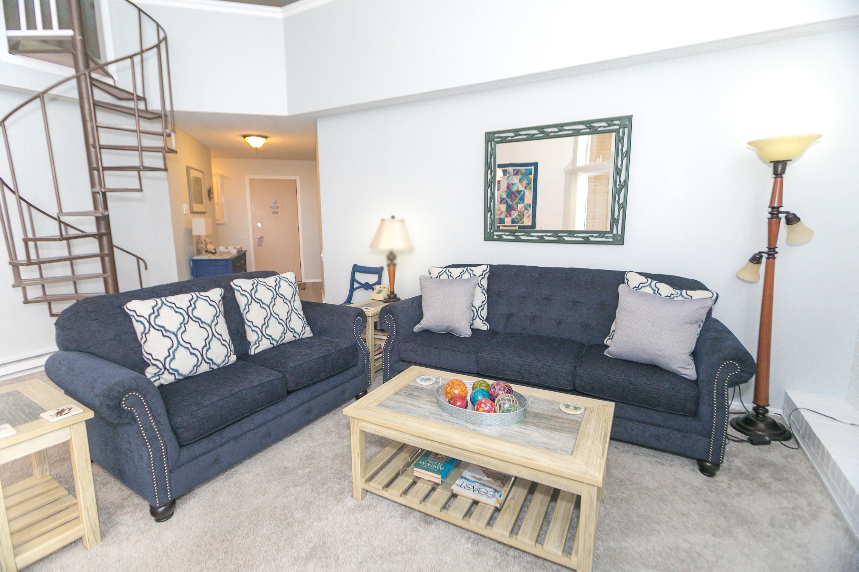 A living room with a dark blue sofa, two patterned throw pillows, a coffee table with a bowl of colorful balls, a lamp, and a mirror on the wall.