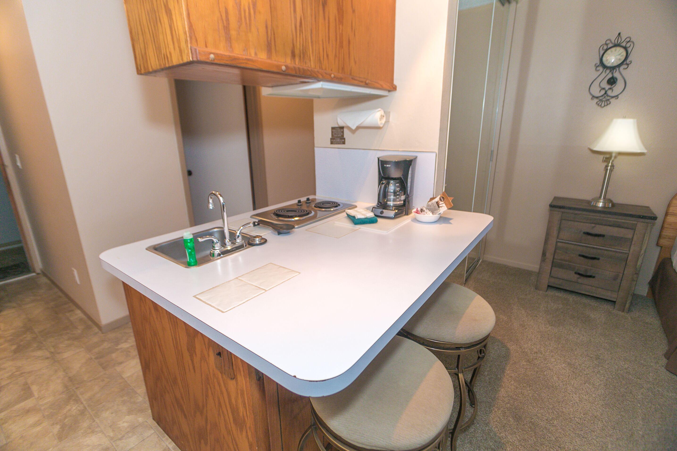 A compact kitchen with a sink, stove, and coffee maker. There are two stools and a small cabinet.