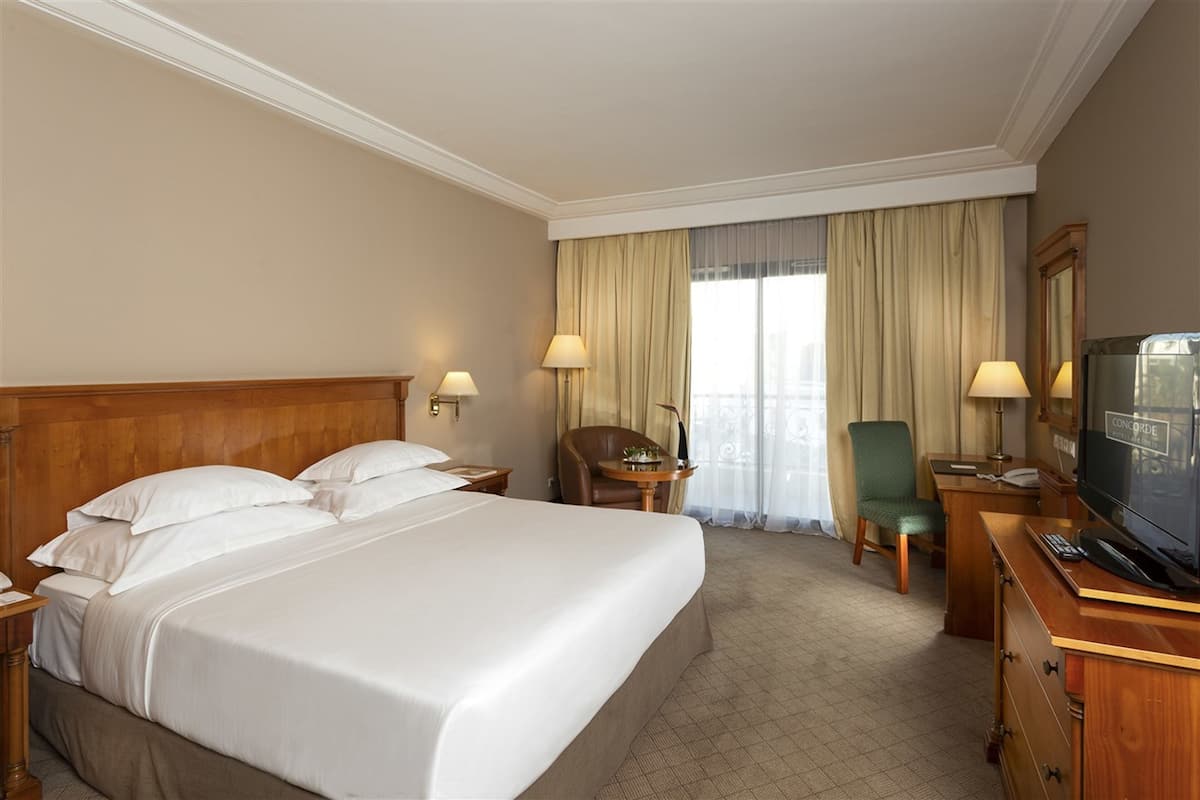 Une chambre d’hôtel avec un grand lit, deux tables de chevet avec des lampes, un bureau avec une chaise, une télévision et une fenêtre avec des rideaux.