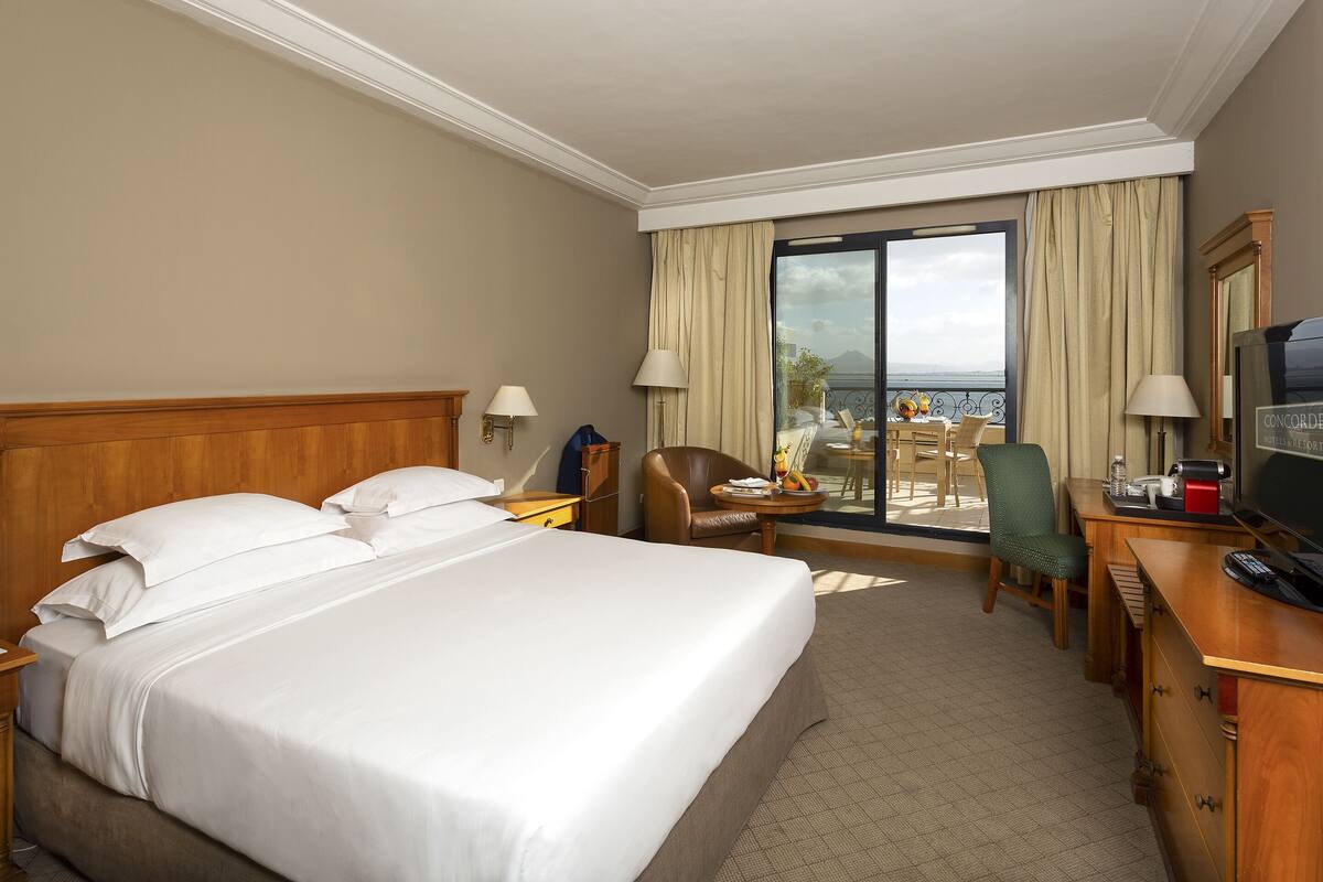 Une chambre d’hôtel avec un grand lit, un bureau, une chaise, une télévision et une vue sur l’océan par une grande fenêtre.